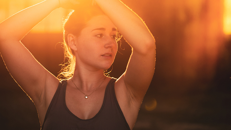 Eine Frau im Top steht im Sonnenlicht und hat die Hände auf den Kopf gelegt.