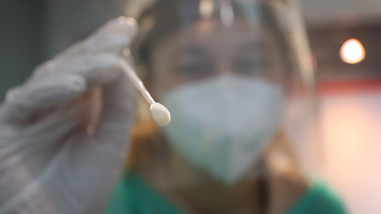 Eine medizinische Assistentin nimmt in einer Teststation mit einem Tupfer einen Abstrich für einen PCR-Test. Sie hält den Tupfer in die Kamera; sie trägt ein durchsichtiges Gesichtsvisier; Foto: dpa (23.01.2022)