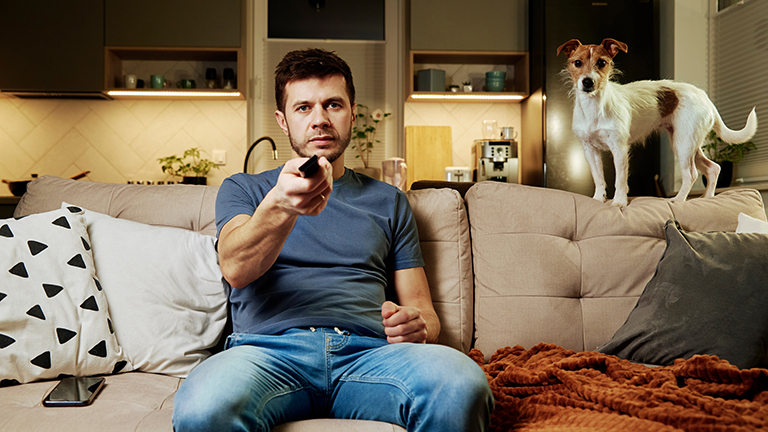 Ein Mann sitzt zu Hause auf einem Sofa und blickt Richtung Kamera; er hält eine Fernbedienung in der Hand. Auf dem Sofa steht ein Hund, der ebenfalls Richtung Kamera blickt.