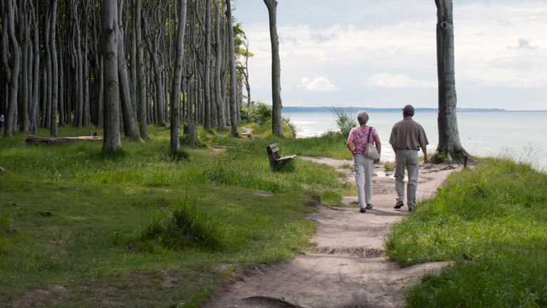Zwei ältere Menschen gehen Hand in Hand am Strand entlang.