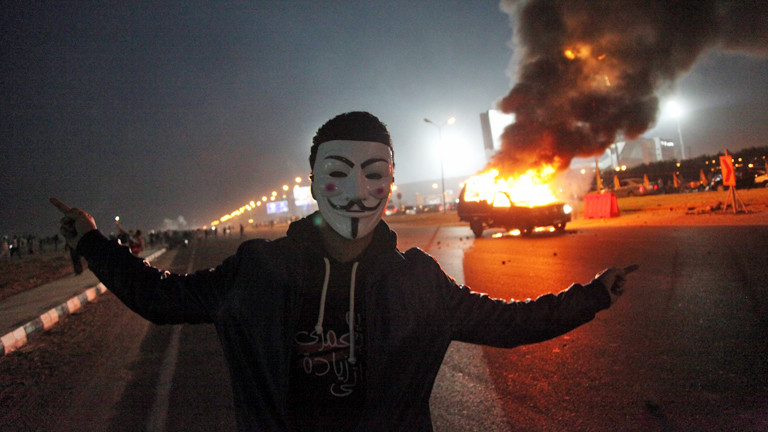 Ein Mann mit Anonymous-Maske steht vor einem brennenden Auto in Ägypten am 08.02.2015.