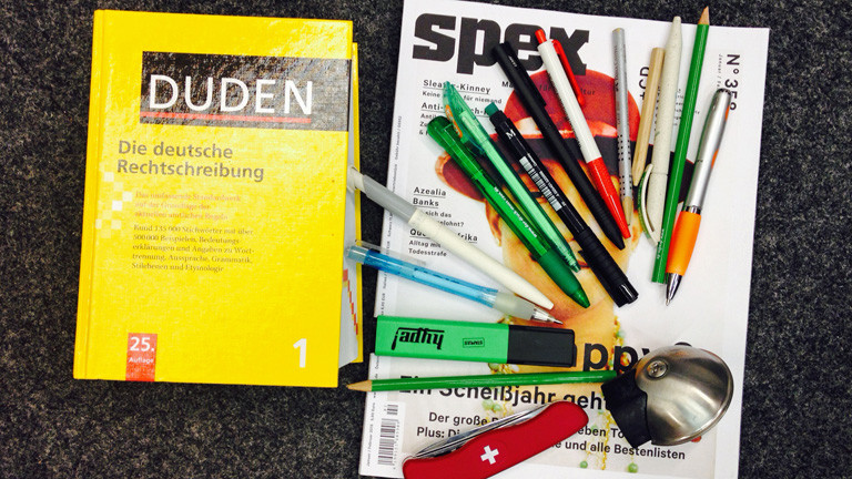 Auf dem Boden liegt ein Duden und ein Magazin, auf dem Stifte, ein Klappmesser und eine Fahrradklingel liegen.