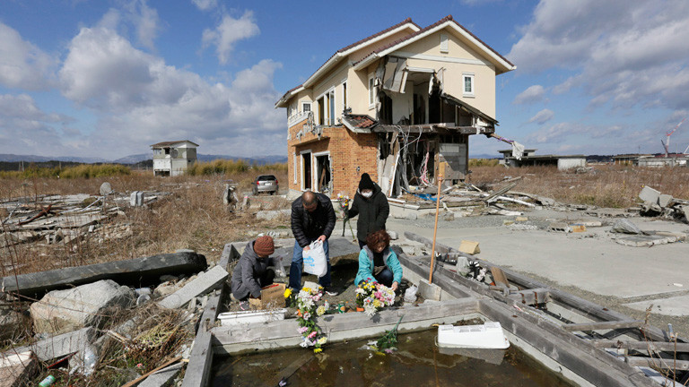Vor einem zerstörten Haus legen vier Personen Blumen nieder. Eine Person trägt einen Mundschutz. Sie gedenken ihren getöteten Eltern in der Stadt Namie in der Präfektur Fukushima (11. März 2015); Bild: dpa