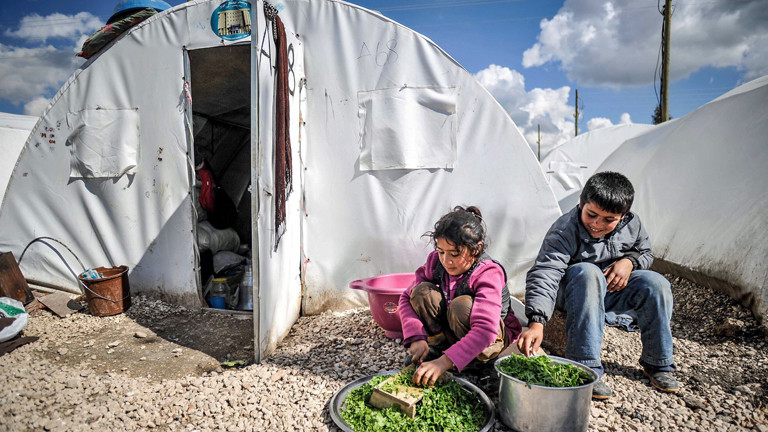 In einem syrischen Flüchtlingslager sitzen zwei Kinder vor einem Zelt und sind mit Lebensmitteln beschäftigt.