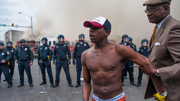 Ein Mann mit nacktem Oberkörper steht vor Polizisten. Er wird von einem älteren Mann zurückgehalten, in der US-Stadt Baltimore am 27.04.2015; Bild: dpa