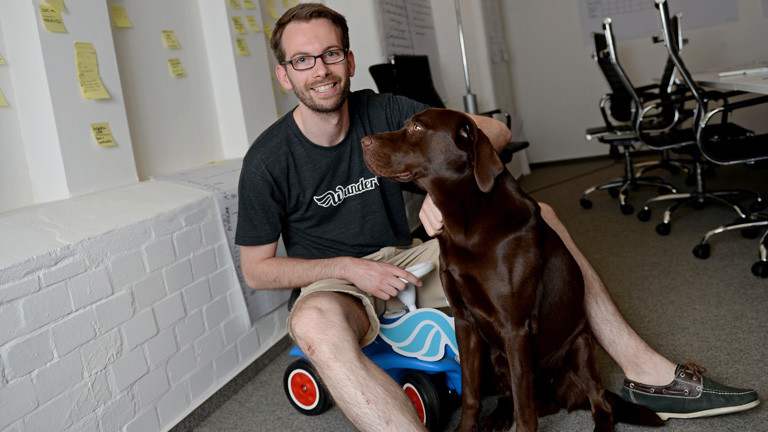 Gunnar Froh, Geschäftsführer der Firma Wundercar, sitzt in einem Büro auf einem Bobby-Car, ein brauner Hund sitzt neben ihm; Bild: dpa