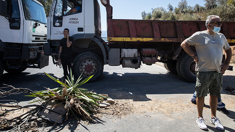 Auf der Insel Lesbos haben Bewohner eine Straßenblockade errichtet; zwei Lkw stehen quer auf der Straße; ein Mann mit Mundschutz steht davor (10.09.2020)