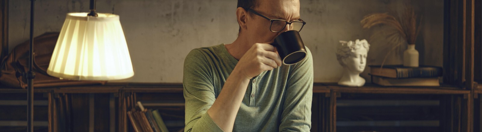 Ein Mann mittleren Alters sitzt an einem Schreibtisch in einem gemütlichen Zimmer und schlürft einen Kräutertee aus einer Tasse. Das sanfte Licht einer Lampe beleuchtet das Buch, das er vor dem Schlafengehen liest