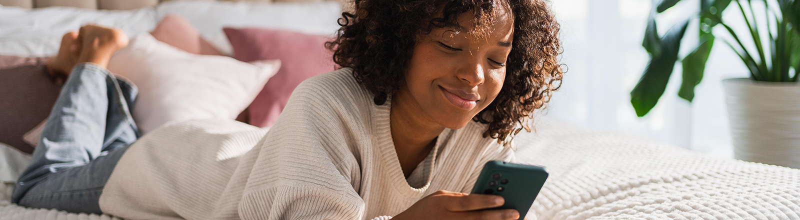 Foto einer jungen Frau, die lächelnd auf dem Bett liegt und auf ihrem Smartphone tippt