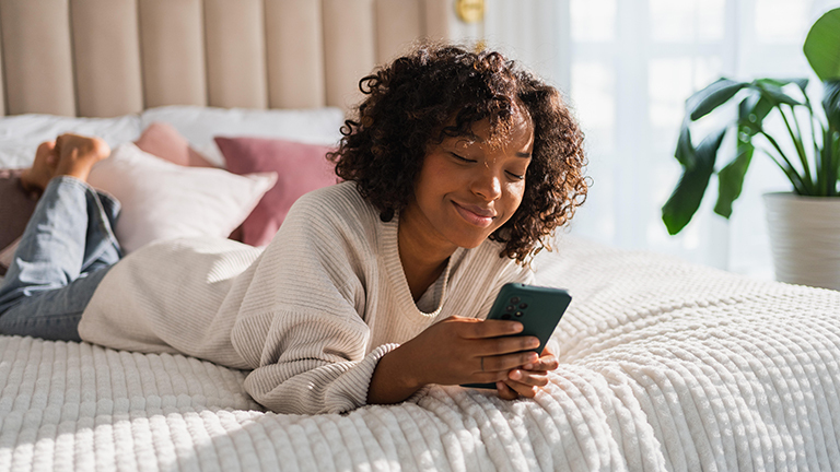 Foto einer jungen Frau, die lächelnd auf dem Bett liegt und auf ihrem Smartphone tippt