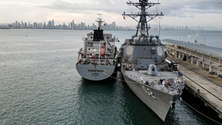 Zwei Schiffe liegen nebeneinander an einem Pier. Eines ist ein Kriegsschiff, das andere ein Versorgungsschiff. Im Hintergrund ist die Skyline einer Stadt zu sehen.