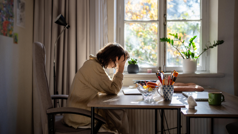 Eine offensichtlich verschlafene, halbwache Frau sitzt an einem Tisch und stützt den Kopf auf ihre rechte Hand