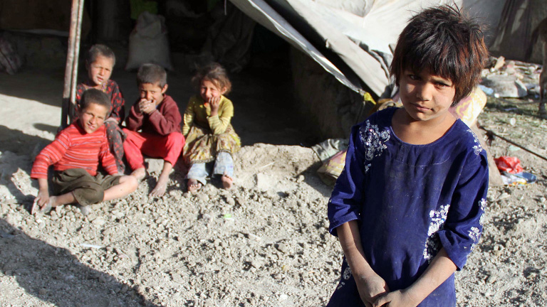 Ein afghanisches Flüchtlingskind steht vor einem Zelt, dort sitzen vier weitere Kinder.