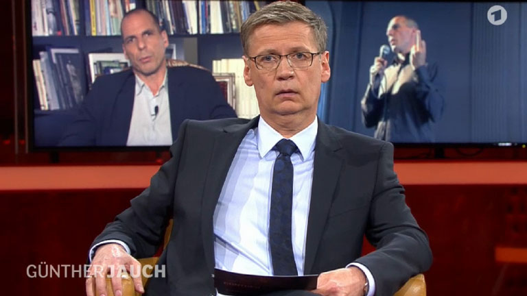 Der Screenshot aus der ARD-Sendung "Günther Jauch", die am 15.03.2015 ausgestrahlt wurde, zeigt den Moderator Günther Jauch; hinter ihm ist auf zwei Monitoren der griechische Finanzminister Gianis Varoufakis zu sehen, links live und rechts in einem Video von 2013 den Stinkefinger zeigend.