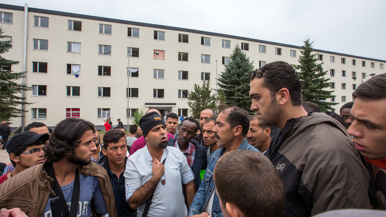 Flüchtlinge stehen am 20.08.2015 vor der teilweise zerstörten Erstaufnahmeeinrichtung in Suhl (Thüringen) und sprechen über die Krawalle der vergangenen Stunden.