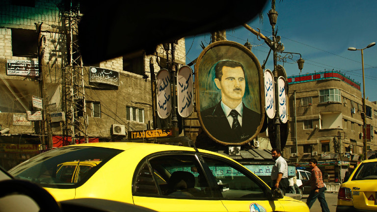 Eine dicht befahrene Straße in Damaskus. Im Hintergrund hängt ein großes Porträt von Assad.