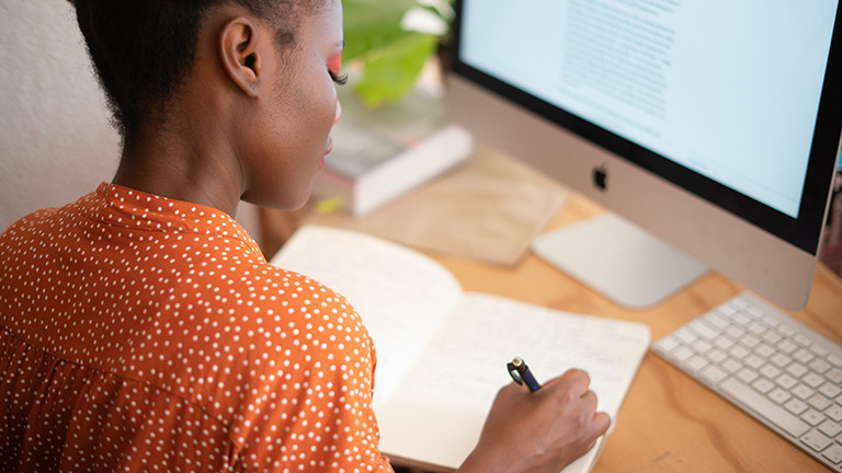 Eine junge Frau sitzt vor einem Computerbildschirm und schreibt in ihr Notizblock