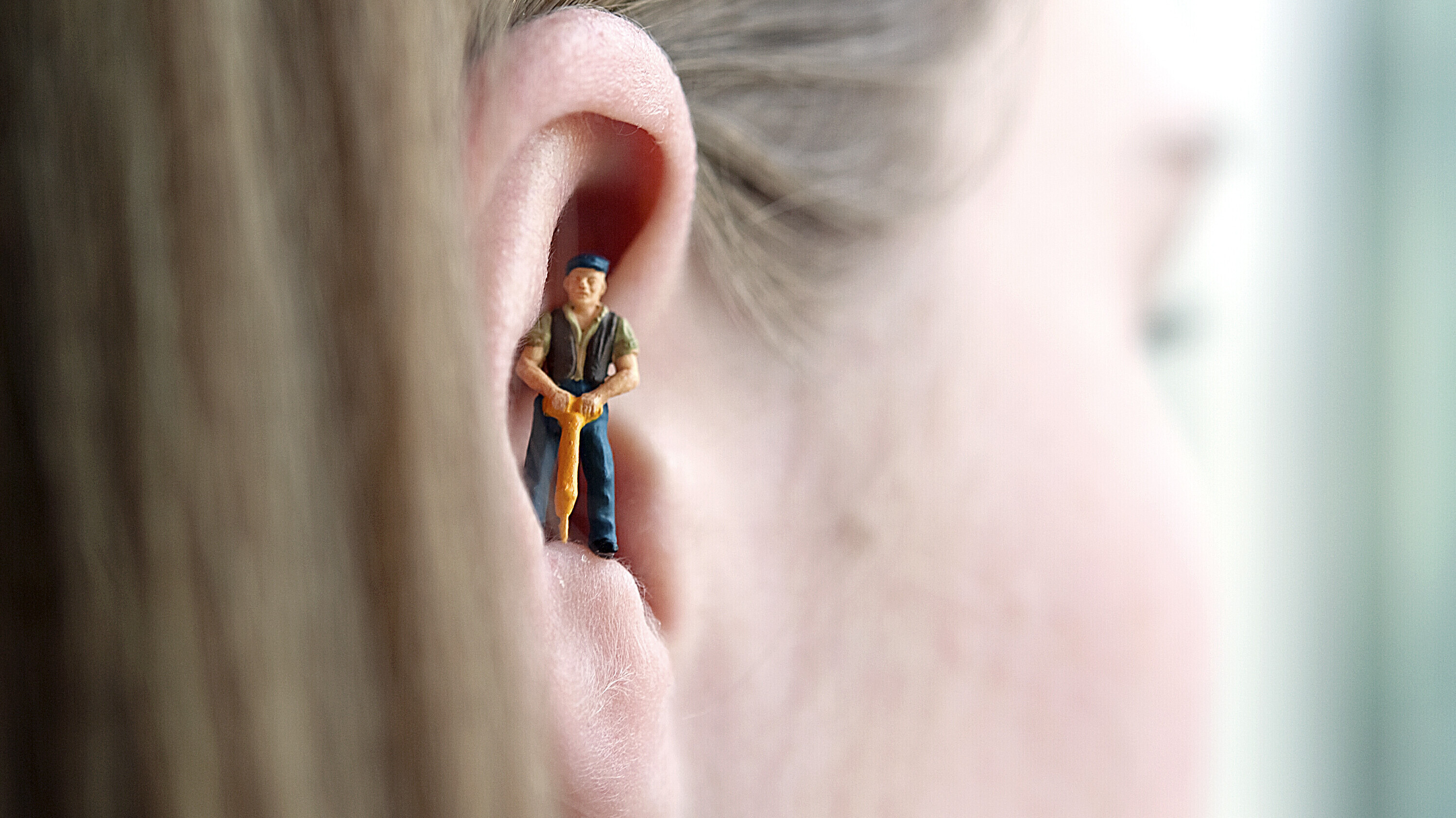 Eine Miniaturfigur eines Bauarbeiters mit Schlagbohrer steht im Ohr einer Frau