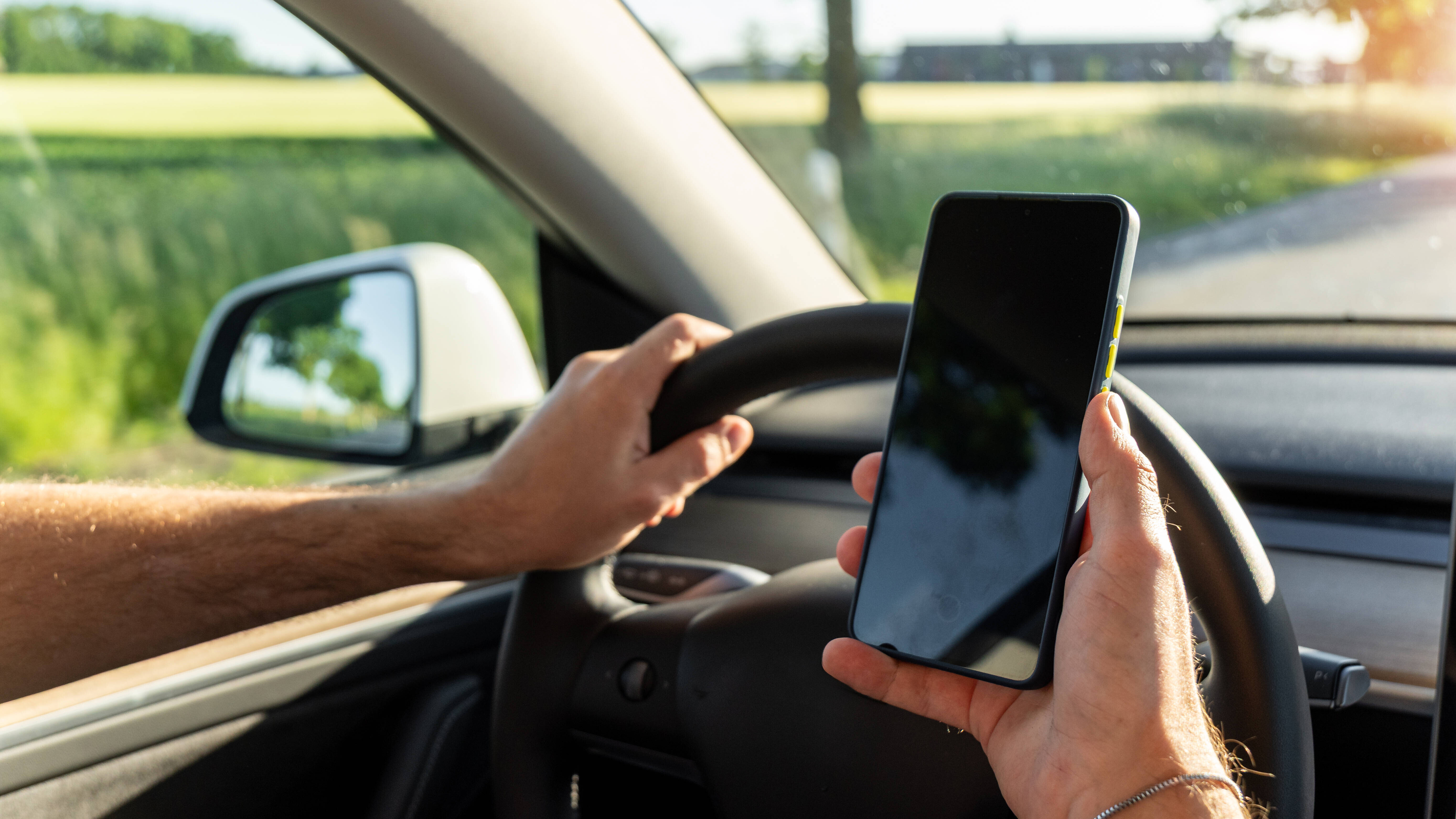 Ein Mann sitzt am Steuer eines fahrenden Autos und hält ein Smartphone in der Hand.