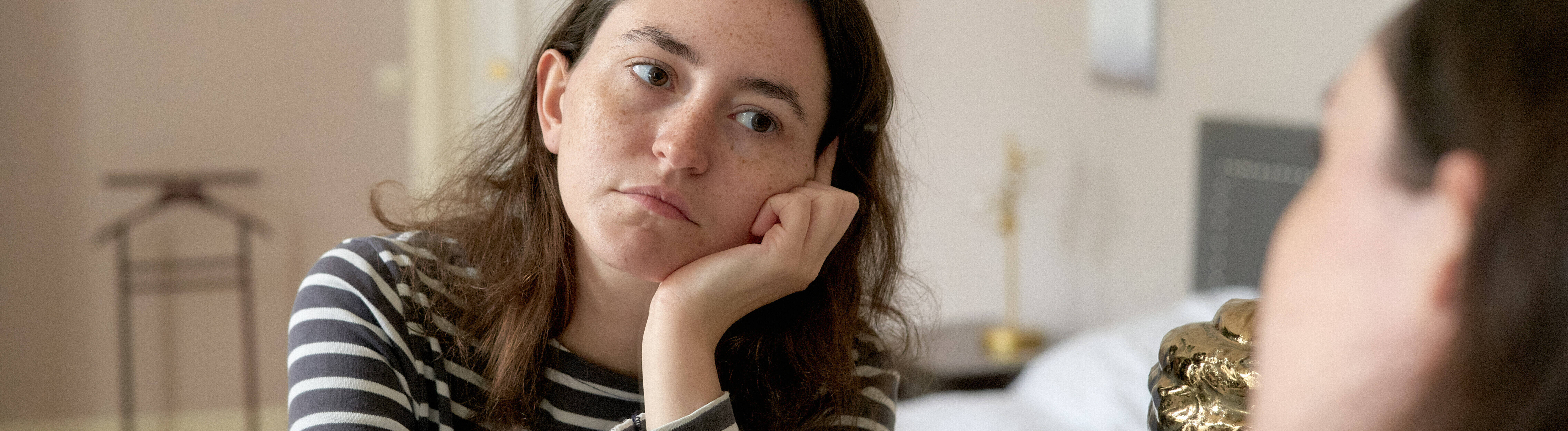 Eine junge Frau in einem gestreiften Oberteil sieht sich selbst in einem Spiegel an. Sie wirkt gelangweilt oder traurig.