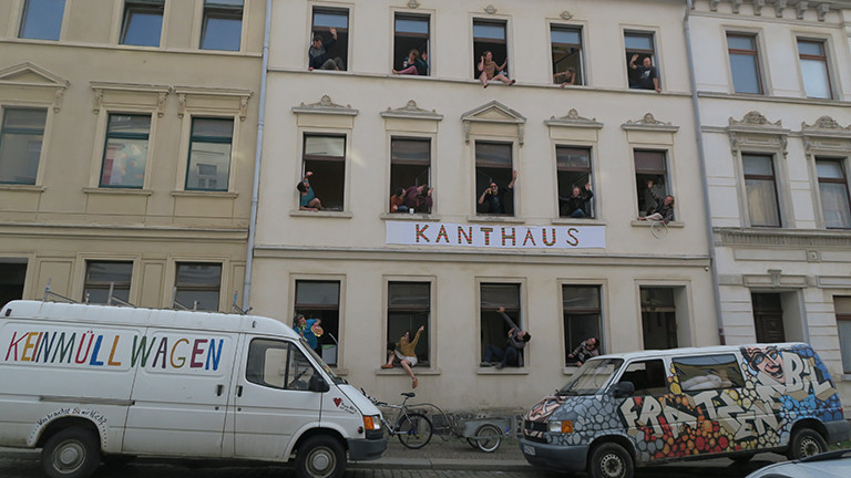 Aus jedem Fenster eines Hauses winken Menschen, auf einem Schild steht Kanthaus.