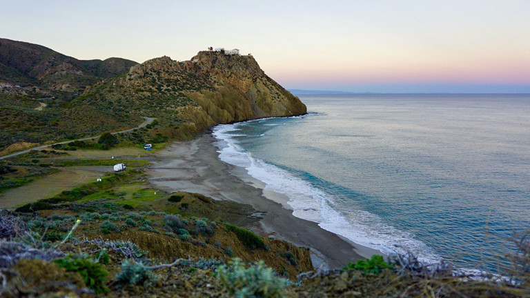 Ein weißer Camper steht klein und verloren in einer malerischen Küstenlandschaft in Spanien