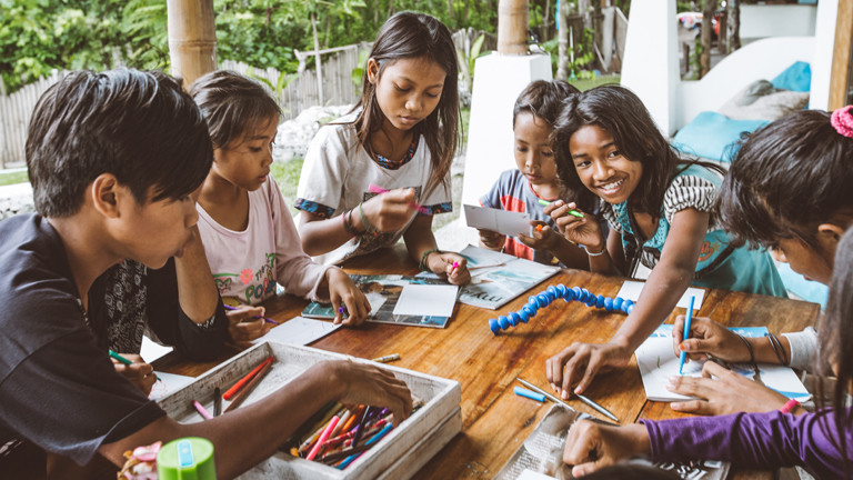 Indonesische Kinder spielen mit Malstiften an einem Tisch.