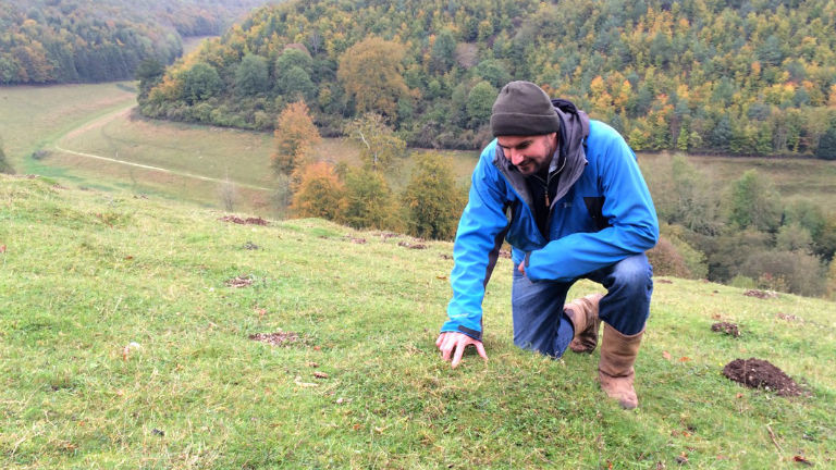 Ein Mann in einer Outdoorjacke zeigt auf eine Stelle im Gras.