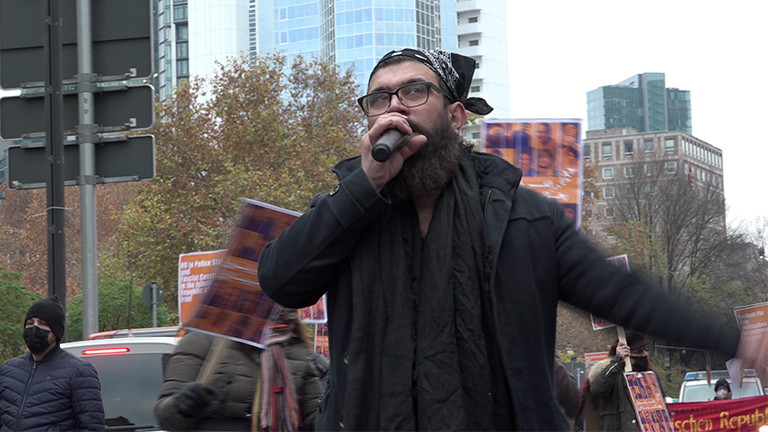 Ein Mann mit Bart und Kopftuch spricht bei einer Demo in ein Mikrofon