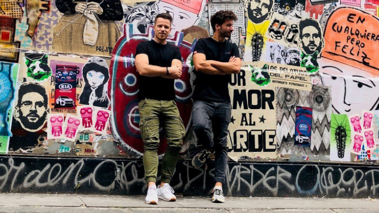 Foto von den zwei Gesprächspartnern Adrian und Christoph, zwei Männern, die vor einer Wand voller bunter Graffiti stehen