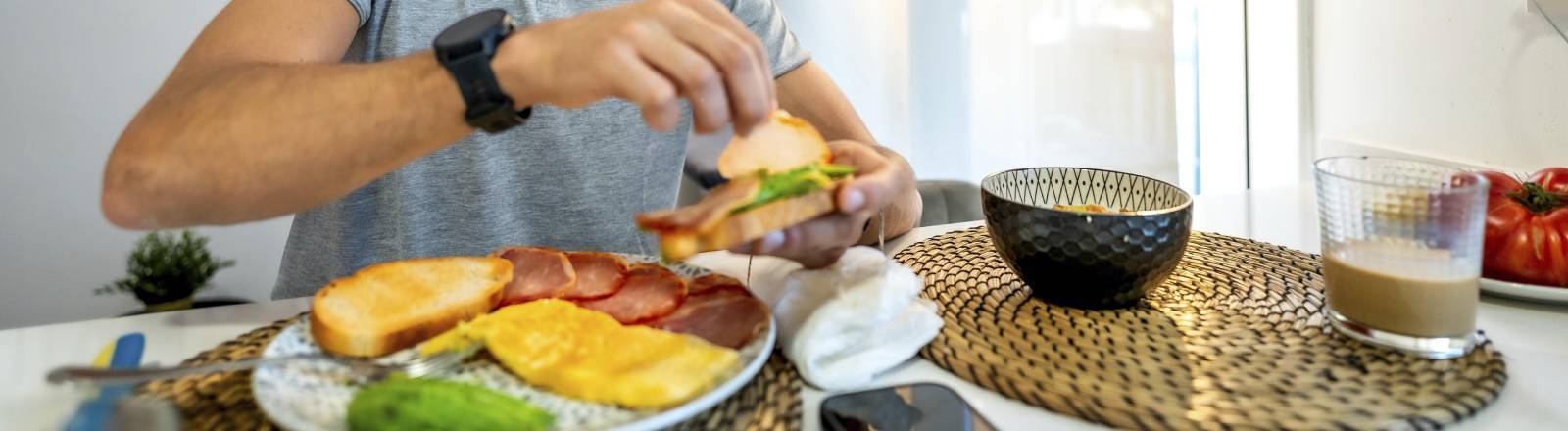 Ein Mann sitzt in der Küche am Frühstückstisch. Vor ihm stehen verschiedene Lebensmittel wie Obst, Brot, Müsli. Zu trinken hat er einen Milchkaffee. Der Mann belegt sich gerade ein Brot. Sein Handy liegt neben dem Teller.