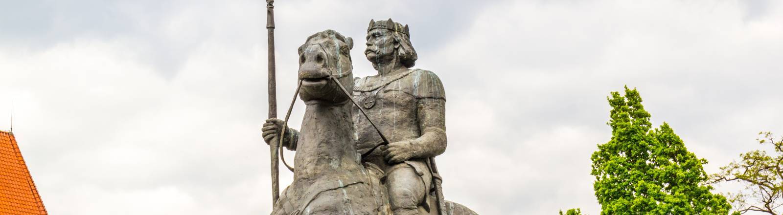 Bronzenes Reiterstandbild von Bolesław I. (dem Tapferen) vor einem bewölkten Himmel, flankiert von grünen Bäumen und dem Dach eines Backsteingebäudes.