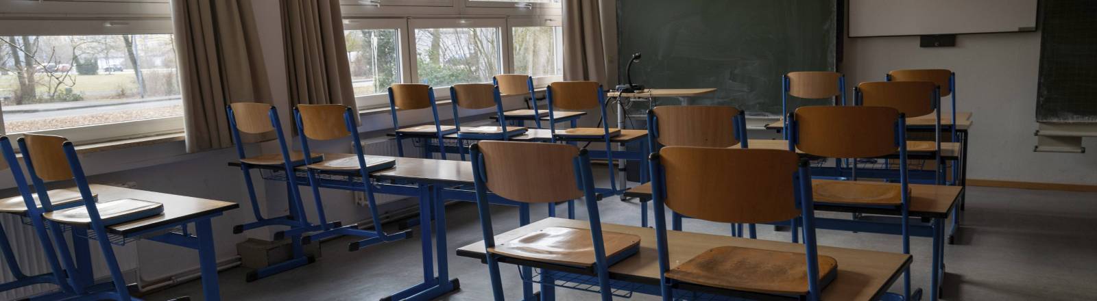 Ein leerer, tageslichtdurchfluteter Klassenraum mit hölzernen Schultischen und Stühlen mit blauen Metallbeinen. Im Hintergrund befinden sich eine Kreidetafel und eine weiße Projektionsfläche an der Wand.