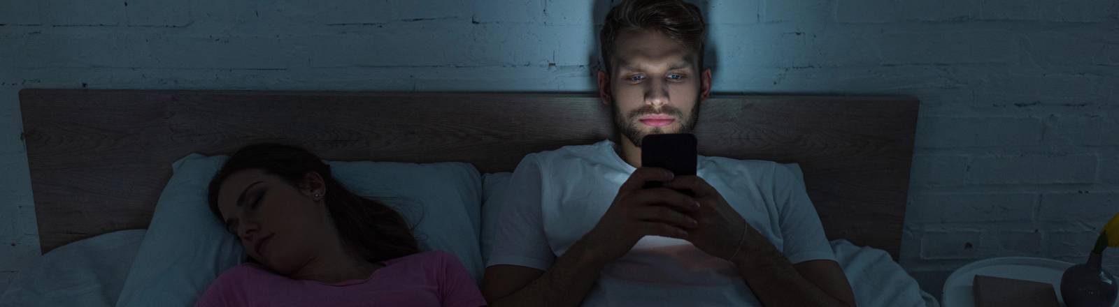 Ein Pärchen liegt im Bett. Der Mann kann nicht schlafen und schaut auf sein Handy. Das Smartphone-Bildschirmlicht hält ihn weiter vom Schlafen ab.