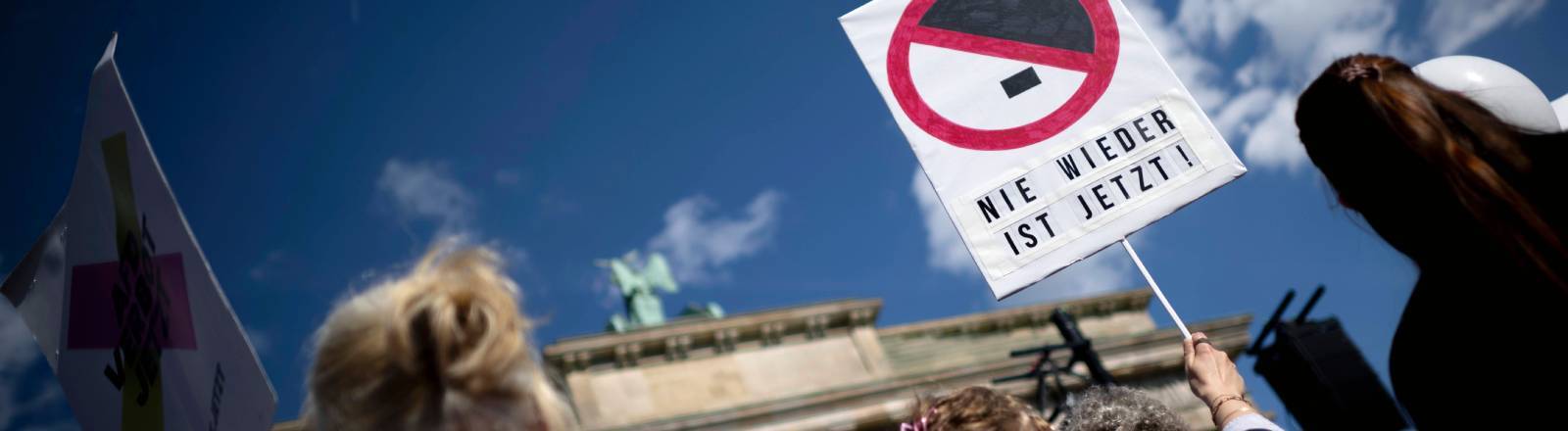 Demonstration gegen Rechtsextremismus vor dem Brandenburger Tor in Berlin; im Fokus hält eine Person ein Schild mit der Aufschrift "NIE WIEDER IST JETZT!" unter einem durchgestrichenen Hitler-Schnitt.