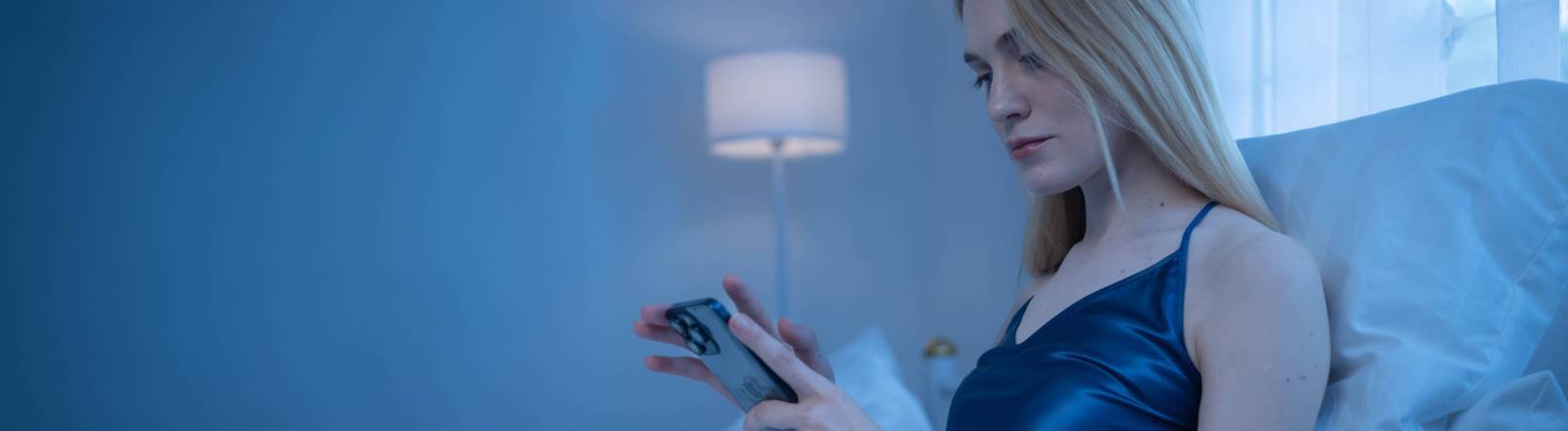 Eine blonde Frau liegt nachts wach in ihrem Bett und schaut auf das Smartphone.