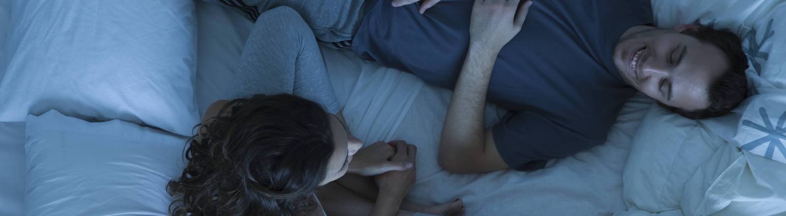 Ein Paar verbringt Zeit im Bett. Frau sitzt. Mann liegt und entspannt.