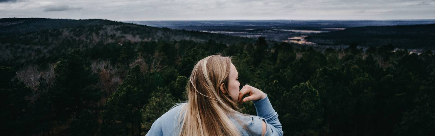 Frau schaut auf Berg- und Meereslandschaft und stützt ihren Kopf dabei auf ihrer Hand ab.