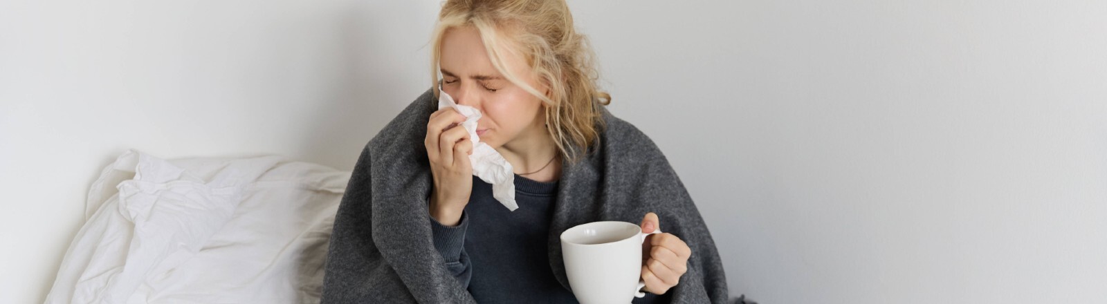 Eine Frau sitzt mit einer Decke um ihre Schultern auf einem Bett. Sie putzt sich mit einem Taschentuch die Nase und hält dabei eine Tasse in der Hand. Vor ihr auf dem Bett liegen ein Laptop und Notizbücher.