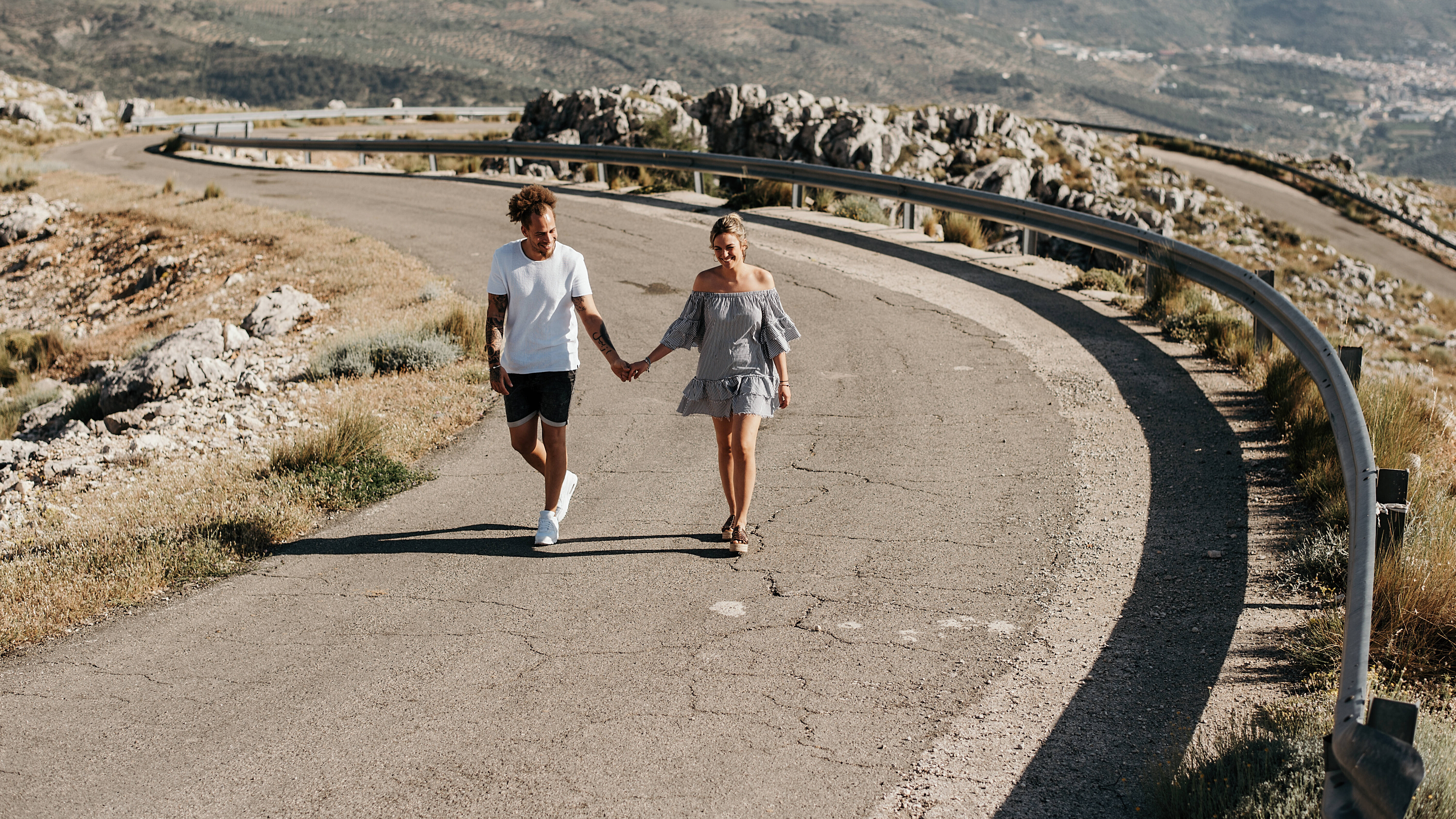 Foto eines Paares, das Hand in Hand auf einer Straße durch eine weiter, felsige Landschaft läuft