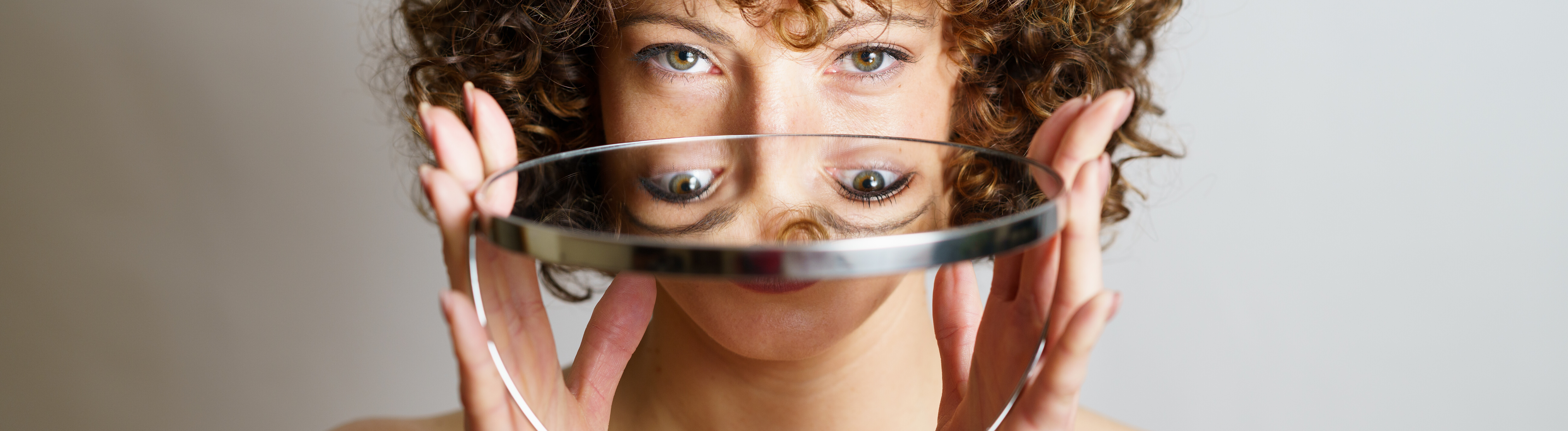 Attraktives junges Model mit lockigem Haar hält einen runden Spiegel in den Händen, in dem sich ihr Gesicht und ihre Augen spiegeln, während sie vor grauem Hintergrund in die Kamera blickt