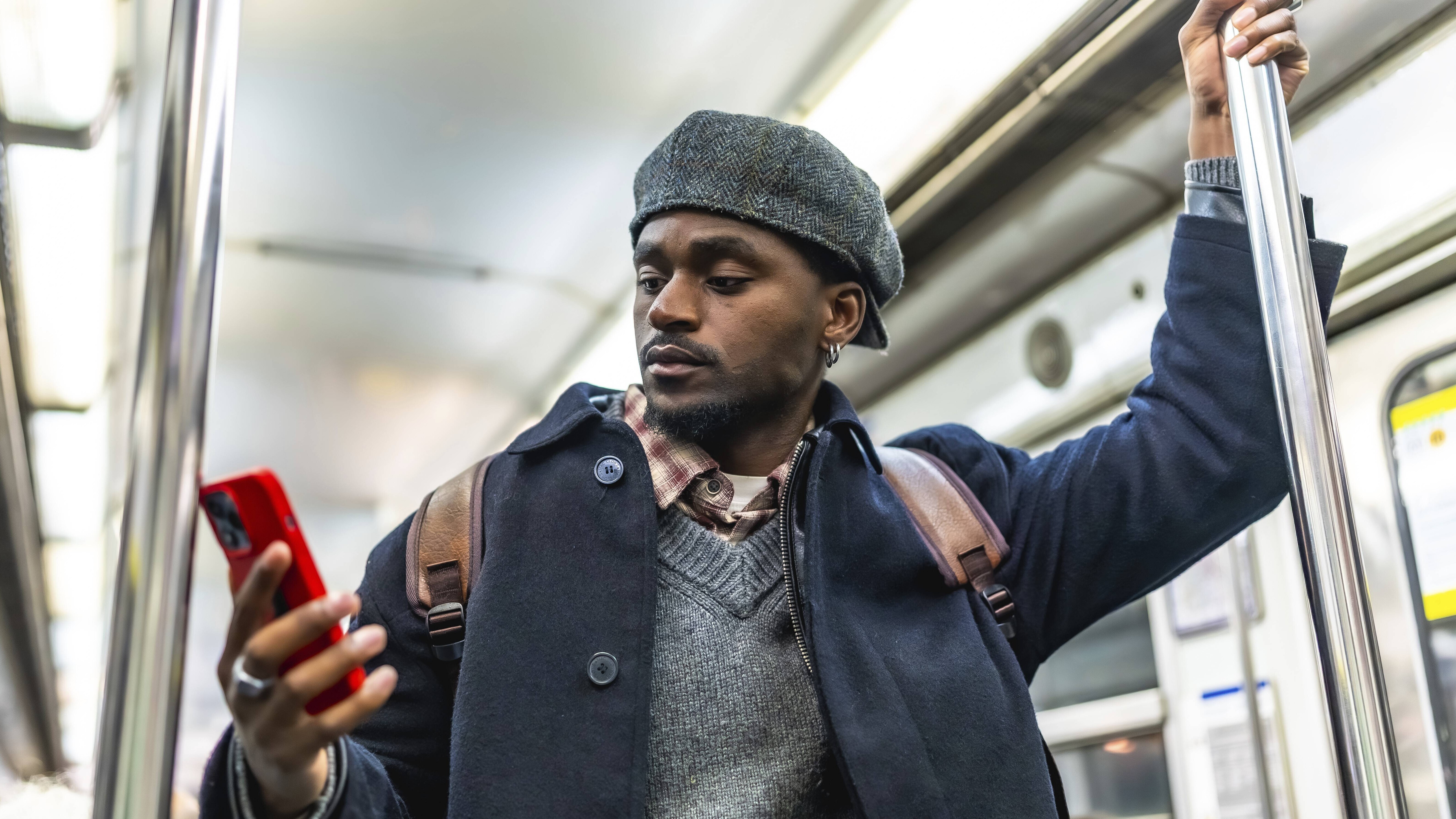 Ein Mann steht in einer U-Bahn. Er hält sich mit einer Hand an einer Stange fest. Mit der anderen Hand hält er sein Handy. Er schaut auf den Handy-Bildschirm.