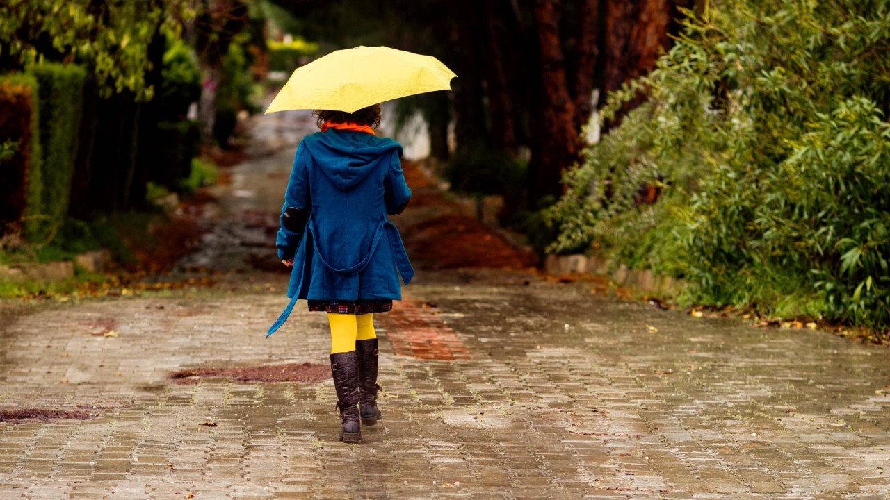 Rückenansicht einer Frau im Mantel, mit gelbem Regenschirm und Gummistiefeln.