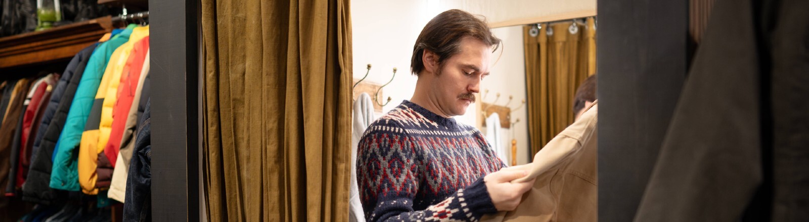 Mann kauft Vintage-Kleidung in einem Second-Hand-Laden und probiert eine Jacke in der Umkleidekabine an.
