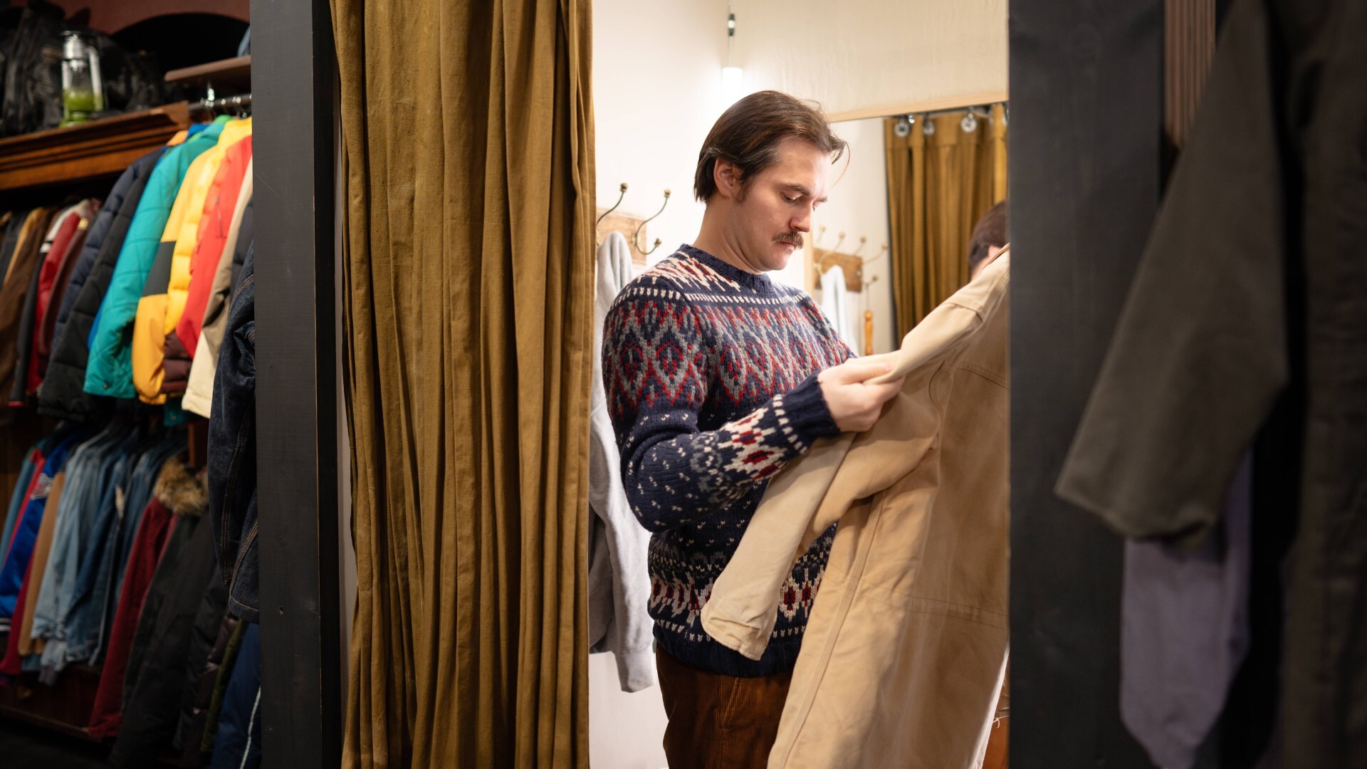 Mann kauft Vintage-Kleidung in einem Second-Hand-Laden und probiert eine Jacke in der Umkleidekabine an.