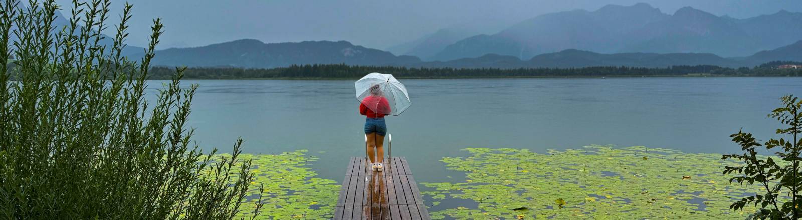 Eine Frau steht auf einem Steg, hält einen Regenschirm in der Hand und blickt auf einen See