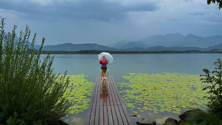 Eine Frau steht auf einem Steg, hält einen Regenschirm in der Hand und blickt auf einen See