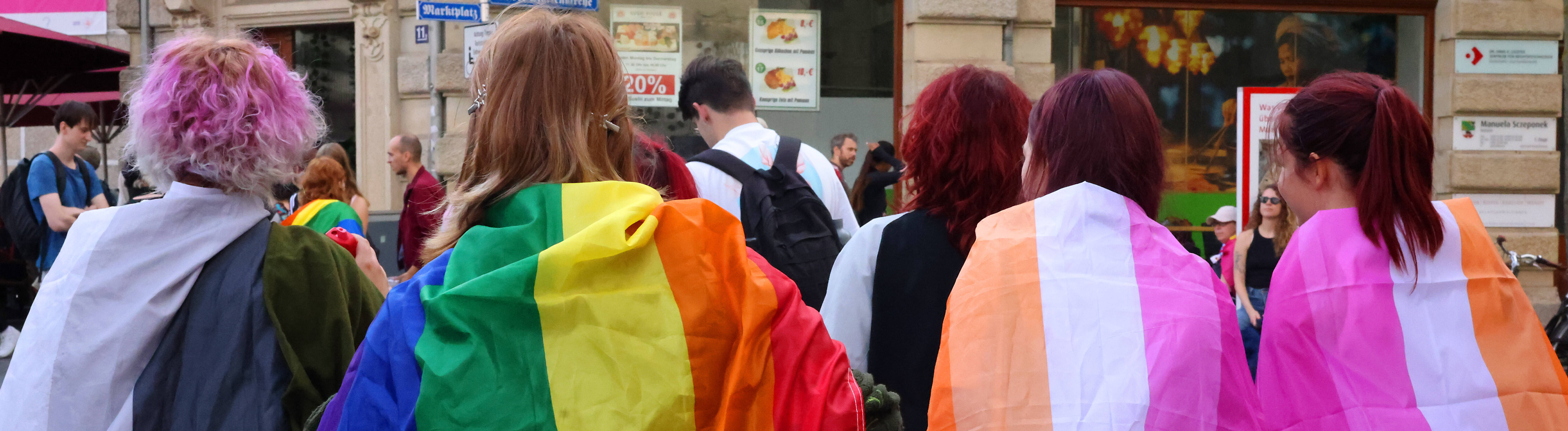 Vier Teilnehmende der CSD-Demonstration von hinten in bunte Flaggen gehüllt (Halle, 12.09.2026)