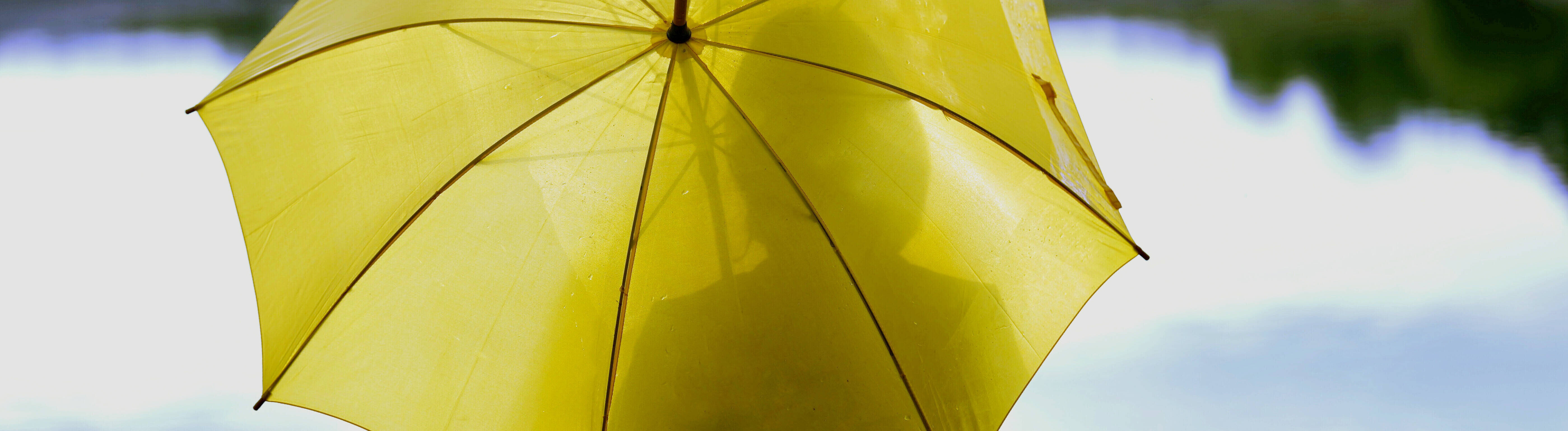 Auf einer Bank an einem See sitzt eine Frau. Sie hat einen großen gelben Regenschirm geöffnet.