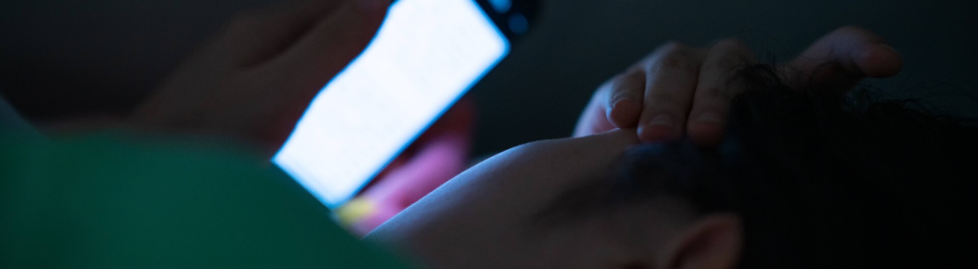 Foto einer jungen Frau, die im Dunkeln im Bett liegt und auf ihr blaues Smartphone-Display guckt